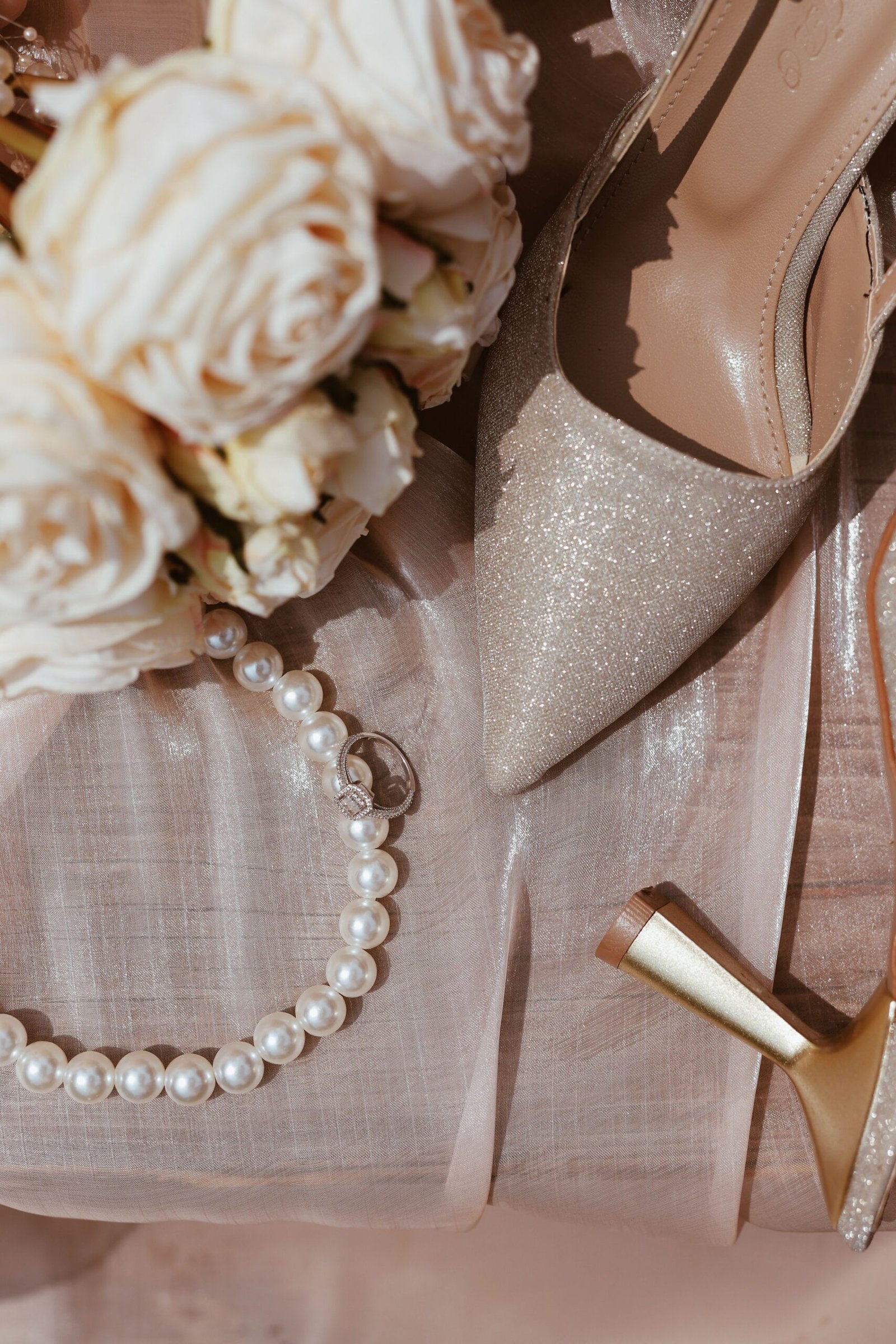 Flat lay of elegant wedding shoes, pearl necklace, and roses on satin fabric.
